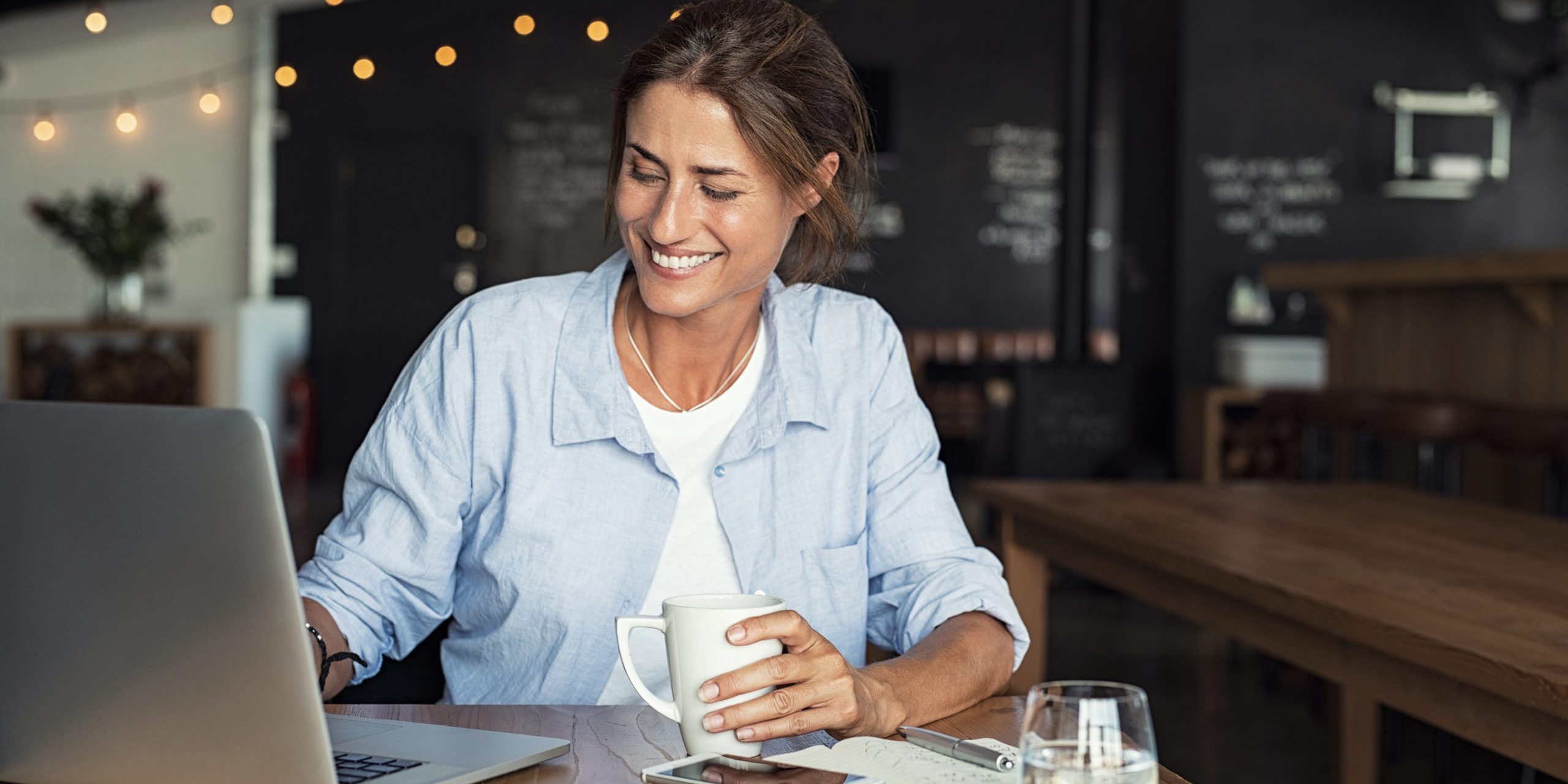Smiling woman sitting in cafeteria holding coffee mug and working on laptop. Businesswoman checking email on laptop. Beautiful middle aged woman smiling and using laptop at cafe while drinking a cup of tea.